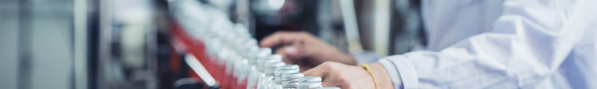A factory worker wearing protective clothing and a mask is overseeing a production line where glass bottles filled with a red liquid are being processed. The setting suggests a controlled environment, possibly in a beverage manufacturing facility.
